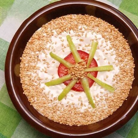 sos tahini w ceramicznej miseczce ozdobiony prażonym sezamem, kawałkiem pomidora i ćwiartkami kiszonych dzikich ogórków